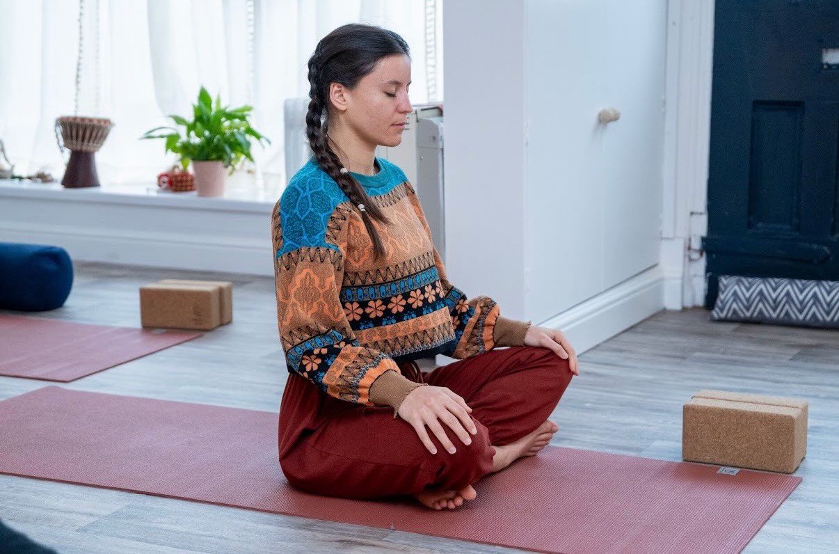 A teacher sitting cross-legged on her mat by the studio window